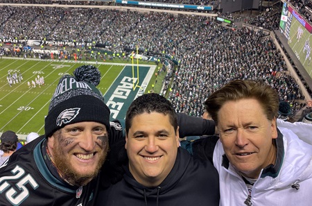 Three men wearing Philadelphia Eagles clothing in crowded stadium with football field in background